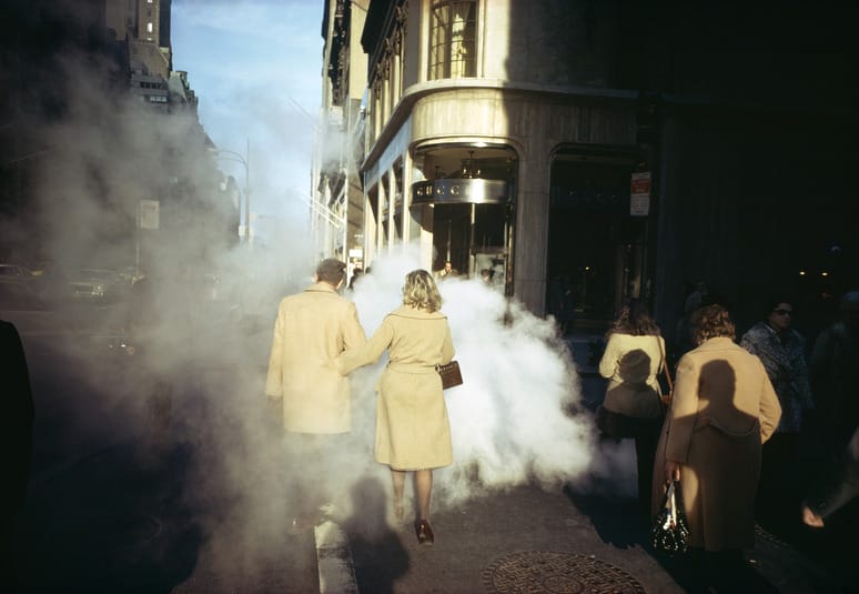 People walking through steam on a city street, wearing camel coats, with sunlight casting shadows on the sidewalk.