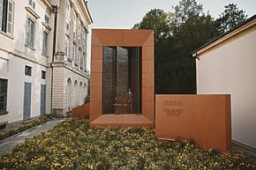 Modern architectural installation with tall orange structure, glass panels, surrounded by blooming yellow flowers and historic buildings.