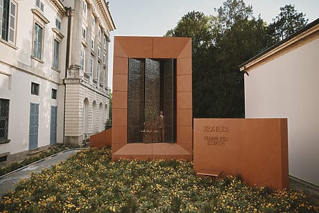 Modern architectural installation with tall orange structure, glass panels, surrounded by blooming yellow flowers and historic buildings.