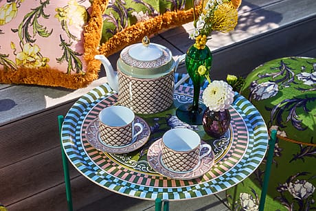 Colorful outdoor table setting with patterned teapot, two teacups, and floral arrangements on a decorative table.