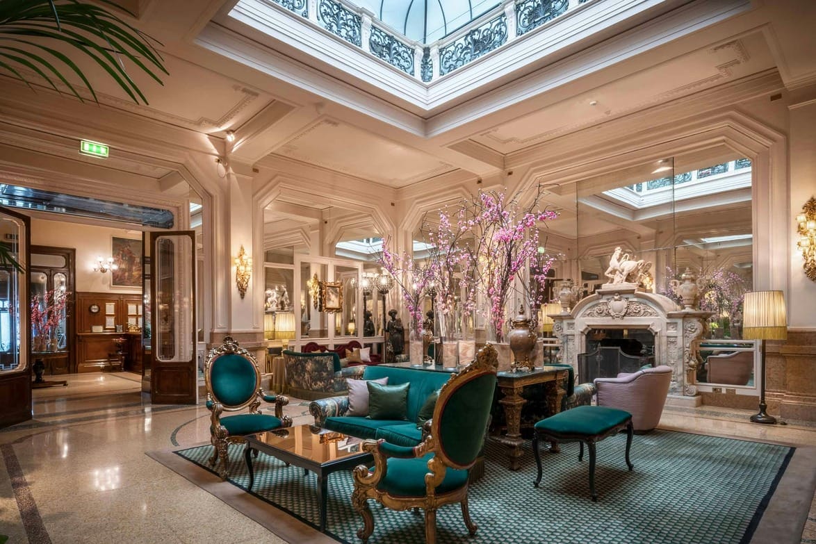 Hotel lobby at Grand Hotel et de Milan, with ornate green chairs, glass ceiling, decorative plants, and art pieces. Bright and inviting ambiance.