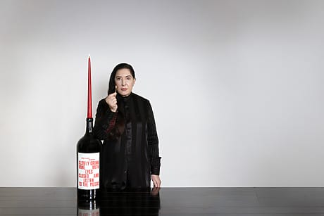 Marina Abramovic in black outfit standing behind a large bottle with red candle on top against a plain background.