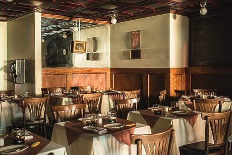 Empty restaurant dining area with neatly arranged tables and chairs, soft lighting, wooden paneling, and framed art on walls.