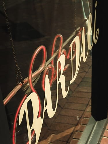 Vintage "Bar Dali" neon sign in a window, reflecting on a brick sidewalk below.