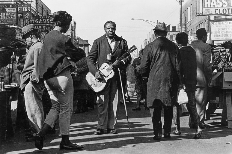 Chicago street musician playing guitar amidst bustling crowd on a busy urban street, surrounded by pedestrians and market activity, photographed by Don Bronstein.