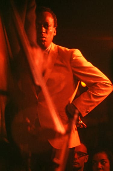 Miles Davis in a dimly lit setting wearing a light-colored suit, captured in a contemplative pose with an intense expression.