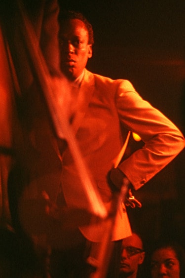 Miles Davis in a dimly lit setting wearing a light-colored suit, captured in a contemplative pose with an intense expression.