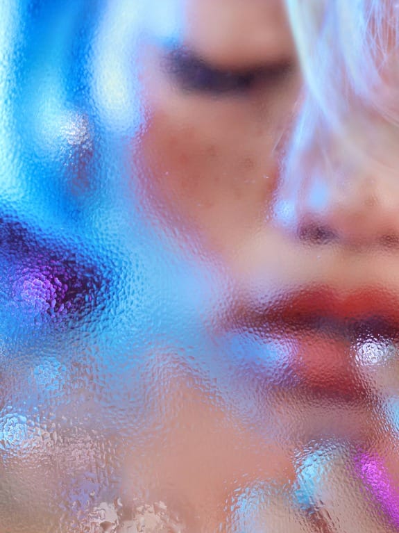 Blurry vibrant close-up of a person's face behind frosted glass with colorful blue and purple lighting effects.