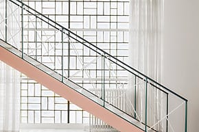 Modern staircase with geometric metal railing and large grid-patterned window in a bright, minimalist interior space.