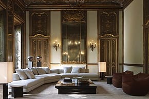 Luxurious living room with ornate gold paneling, large mirror, white sectional sofa, coffee table, and brown chairs.