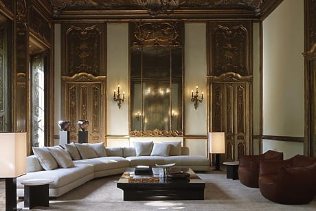 Luxurious living room with ornate gold paneling, large mirror, white sectional sofa, coffee table, and brown chairs.