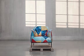 Colorful patchwork armchair on a wooden pallet in a minimalist room with large windows and concrete walls.