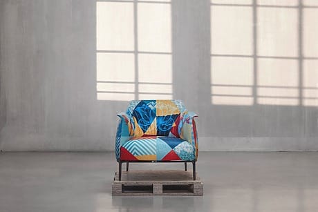 Colorful patchwork armchair on a wooden pallet in a minimalist room with large windows and concrete walls.
