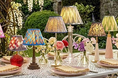 Colorful lamps on an outdoor table set with flowers and checkered napkins against a lush garden backdrop.