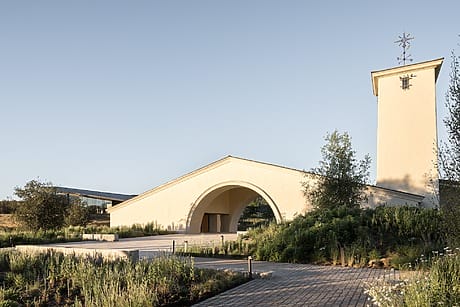 Modern beige chapel with arched entrance, surrounded by lush greenery under a clear blue sky.