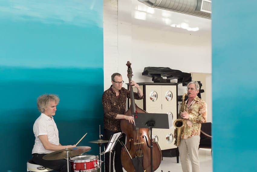 Three musicians playing drums, double bass, and saxophone against a blue wall in a bright room.