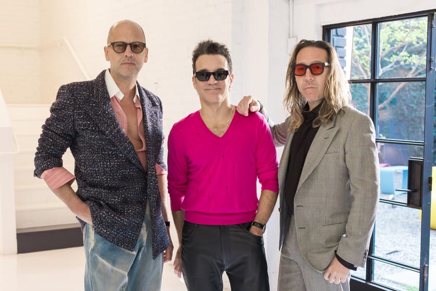 Three men standing stylishly indoors with sunglasses, one in a pink top, the others in patterned and gray suits.