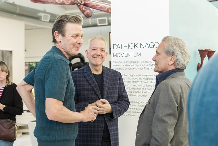 Three men having a conversation at an art exhibition with a display titled "Patrick Naggar: Momentum" in the background.