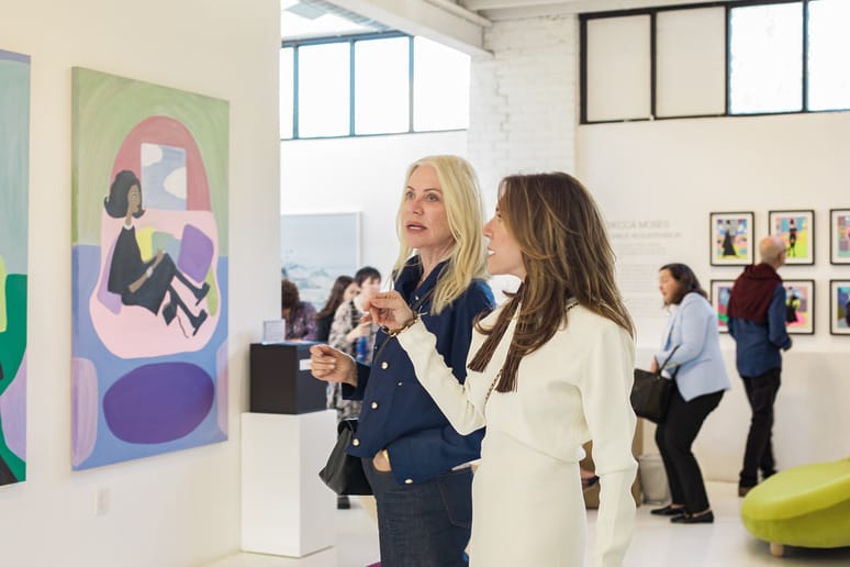 Two women discussing colorful paintings in a bright art gallery with other people in the background.