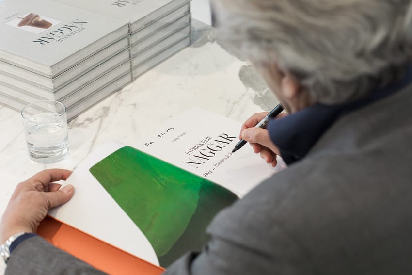 Person signing a book at a table with a glass of water and stacked books nearby.