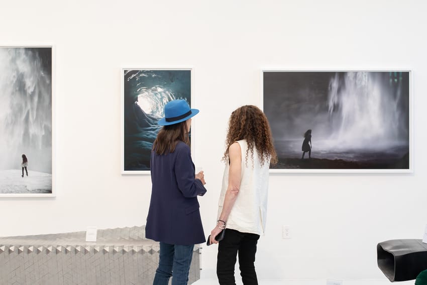 Two people viewing photographs in an art gallery setting, one wearing a blue hat and the other with long curly hair.