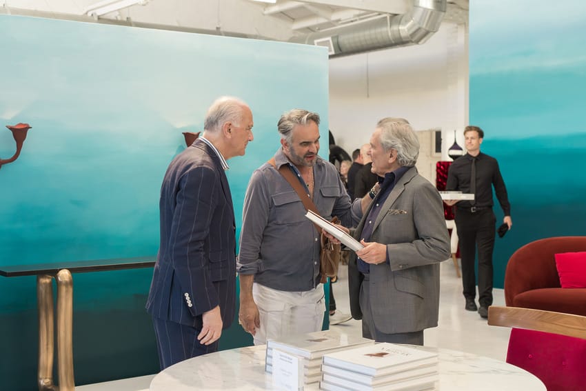 Three men talking and looking at a book in an art gallery with blue and green paintings in the background.