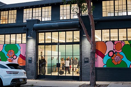 People gather inside a modern building with vibrant floral murals on the exterior, as seen from the street.