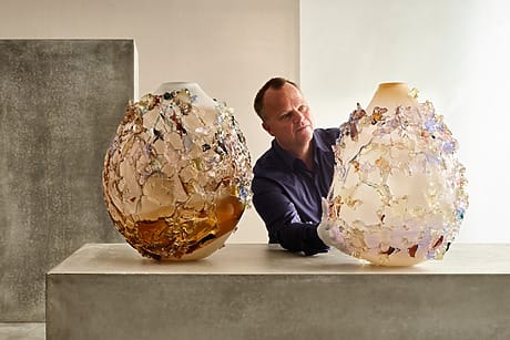Man adjusts two ornate glass vases with floral designs on a table in a minimalistic room setting.
