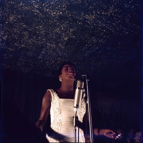 Singer Sarah Vaughn in a white dress sings into a vintage microphone under a textured ceiling in a dimly lit venue, as photographed by Don Bronstein.