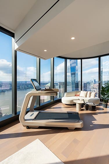 Modern treadmill in spacious room with large windows, overlooking city skyline and river views, next to a stylish white sofa.