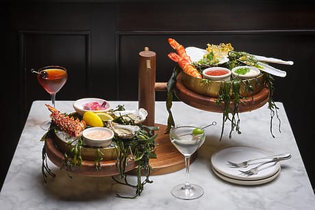 Seafood platter with shrimp, oysters, dipping sauces, lemon garnish, cocktail and martini on a marble table.