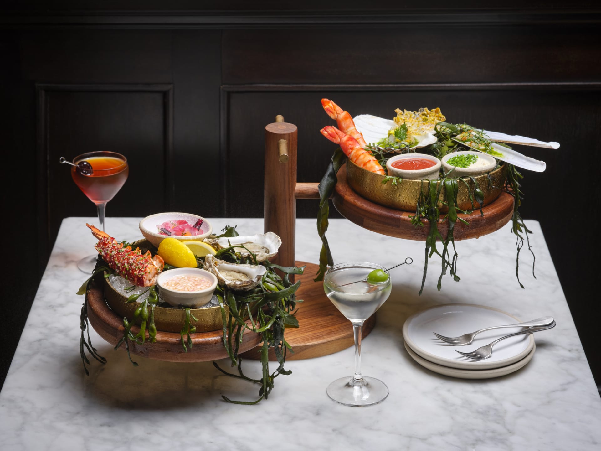 Seafood platter with shrimp, oysters, dipping sauces, lemon garnish, cocktail and martini on a marble table.