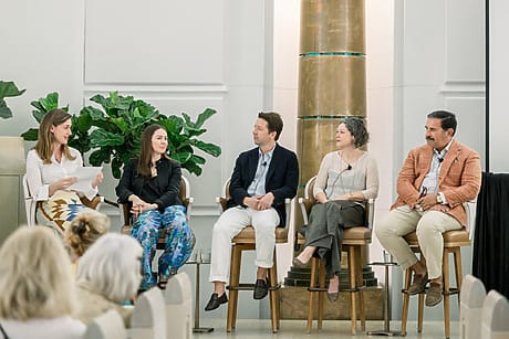 Panel discussion with five people seated in chairs, engaging in conversation at an event, large plant in background.