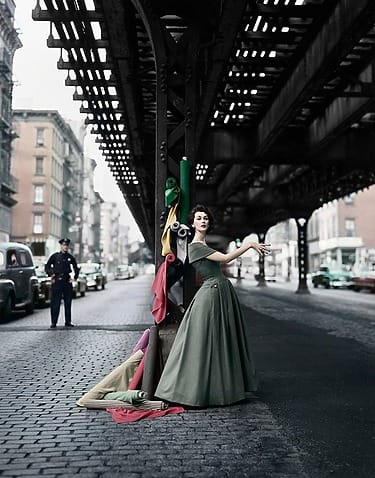 Dovima in a green dress posing under an elevated train track in a city, with colorful fabric rolls beside her, in William Helburn, Dovima Under the El, 1956, at the Park Avenue Armory in the AIPAD show.