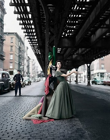 Dovima in a green dress posing under an elevated train track in a city, with colorful fabric rolls beside her, in William Helburn, Dovima Under the El, 1956, at the Park Avenue Armory in the AIPAD show.