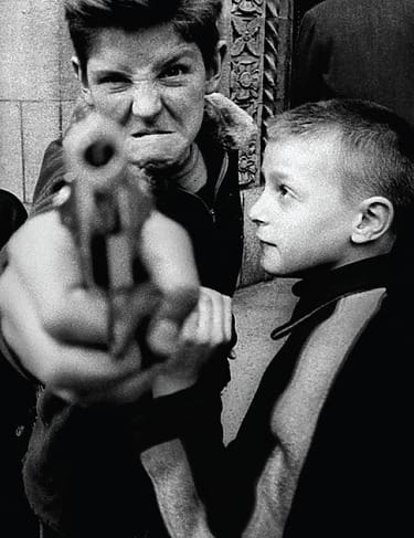 William Klein, Gun 1: Two boys, one points a toy gun at the camera, displaying playful expressions in a candid black-and-white street photo.