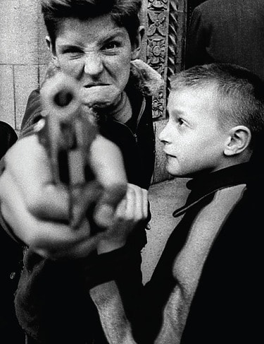 William Klein, Gun 1: Two boys, one points a toy gun at the camera, displaying playful expressions in a candid black-and-white street photo.