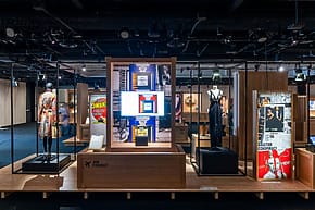 Museum exhibition display with vintage posters, clothing, and informational signage under dim lighting.