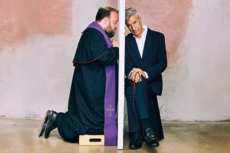 Artist Maurizio Cattelan and a man dressed as a priest pose as if for confession.