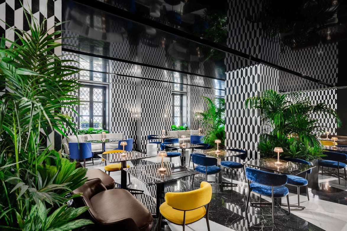 Modern, stylish restaurant interior at the Mandarin Oriental Milan with geometric patterns, blue and yellow chairs, green plants, and reflective surfaces.