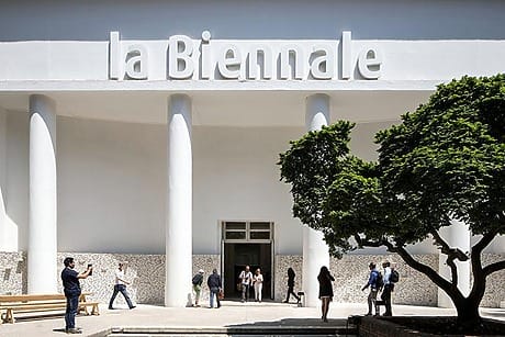 People entering the La Biennale building in Venice with its grand white columns under clear skies, surrounded by trees.