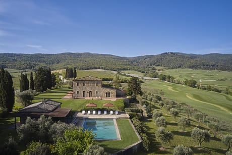 Aerial view of a villa with a pool surrounded by greenery and rolling hills under a clear blue sky.