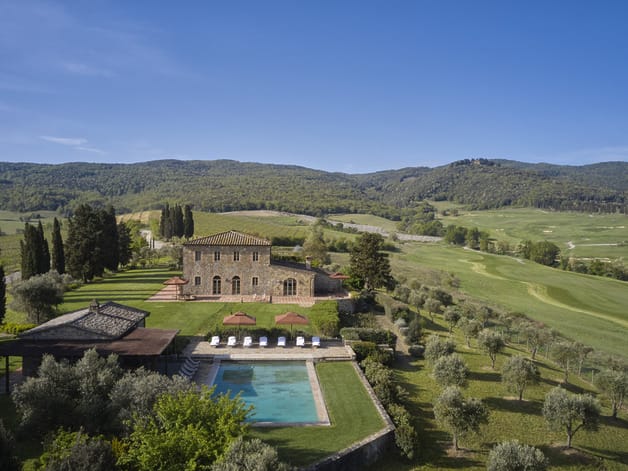 Aerial view of a villa with a pool surrounded by greenery and rolling hills under a clear blue sky.