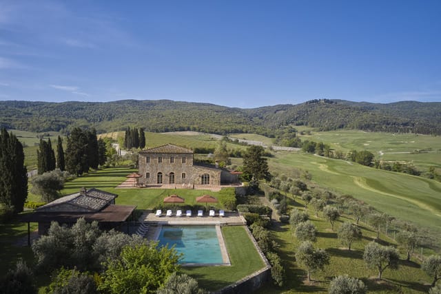 Aerial view of a villa with a pool surrounded by greenery and rolling hills under a clear blue sky.