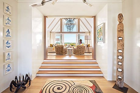 Spacious living room with wicker chairs, large windows, wooden sculpture, abstract wall art, and modern decor.