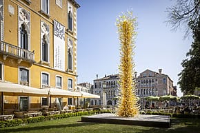 Tall glass sculpture outside a historic yellow building with arched windows and outdoor seating under umbrellas.