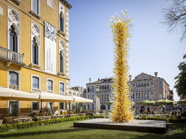 Tall glass sculpture outside a historic yellow building with arched windows and outdoor seating under umbrellas.