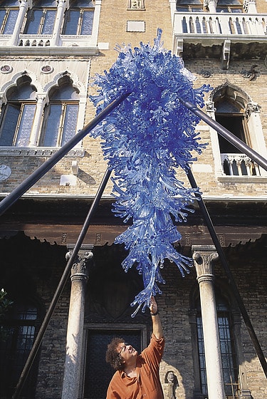 Person reaching towards a large blue glass sculpture suspended above, in front of an ornate building façade.