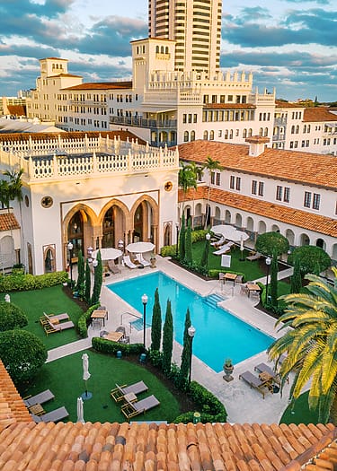Luxury resort with elegant architecture, palm trees, and a central turquoise pool surrounded by lush gardens and lounge chairs.