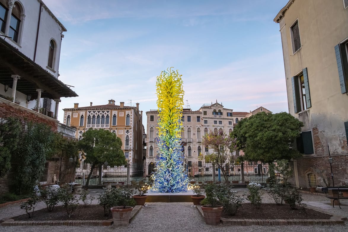 Colorful glass sculpture standing in a courtyard surrounded by historic buildings and trees under a blue sky.